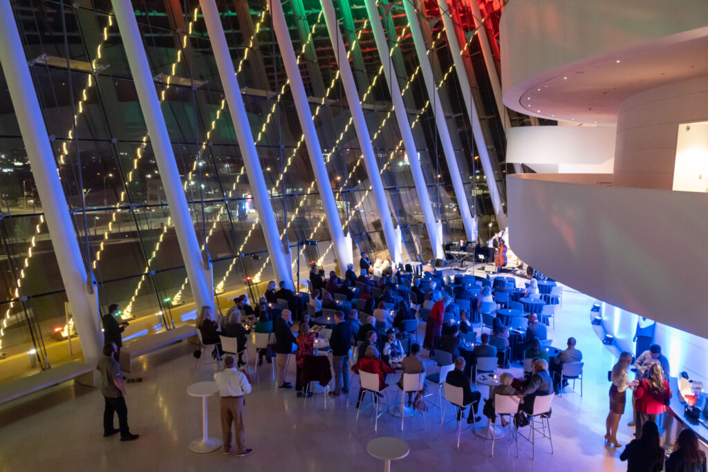 A large group of people gathers in a modern, well-lit indoor space with high ceilings and large windows, featuring a stage area with a musical performance taking place.