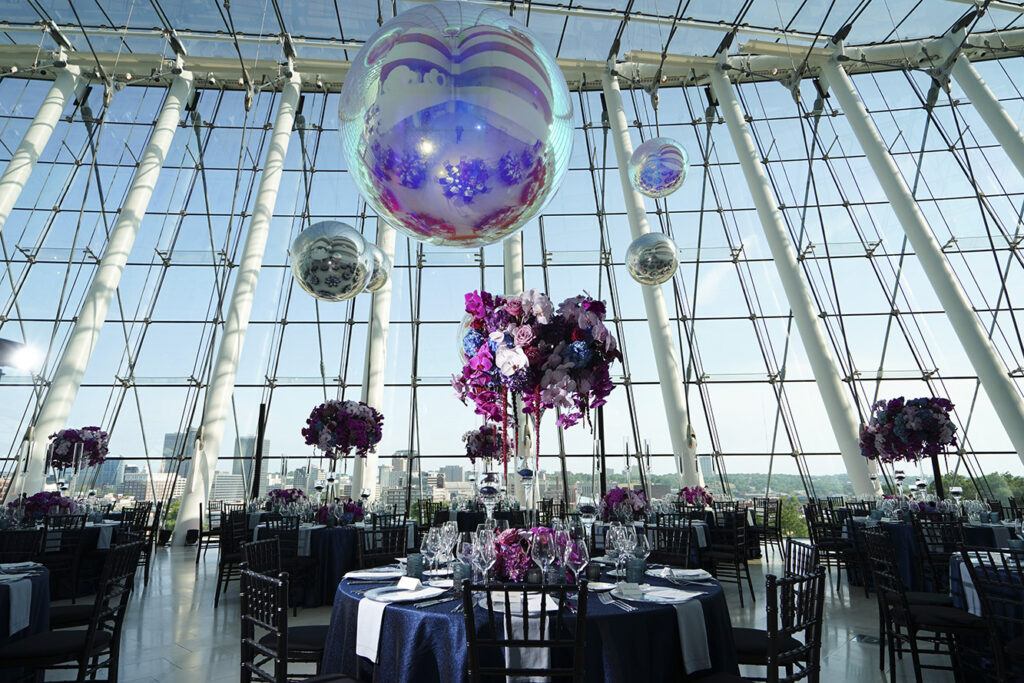 A large room with tall glass windows features round tables with black chairs, purple floral centerpieces, and large decorative balloons hanging from the ceiling. Perfect for events, you can rent this elegant space at the Kauffman Center for an unforgettable experience.