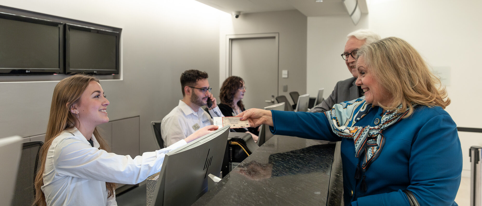 A woman hands a document to a female concierge at the reception desk. Two other receptionists and a man are also present in the background.