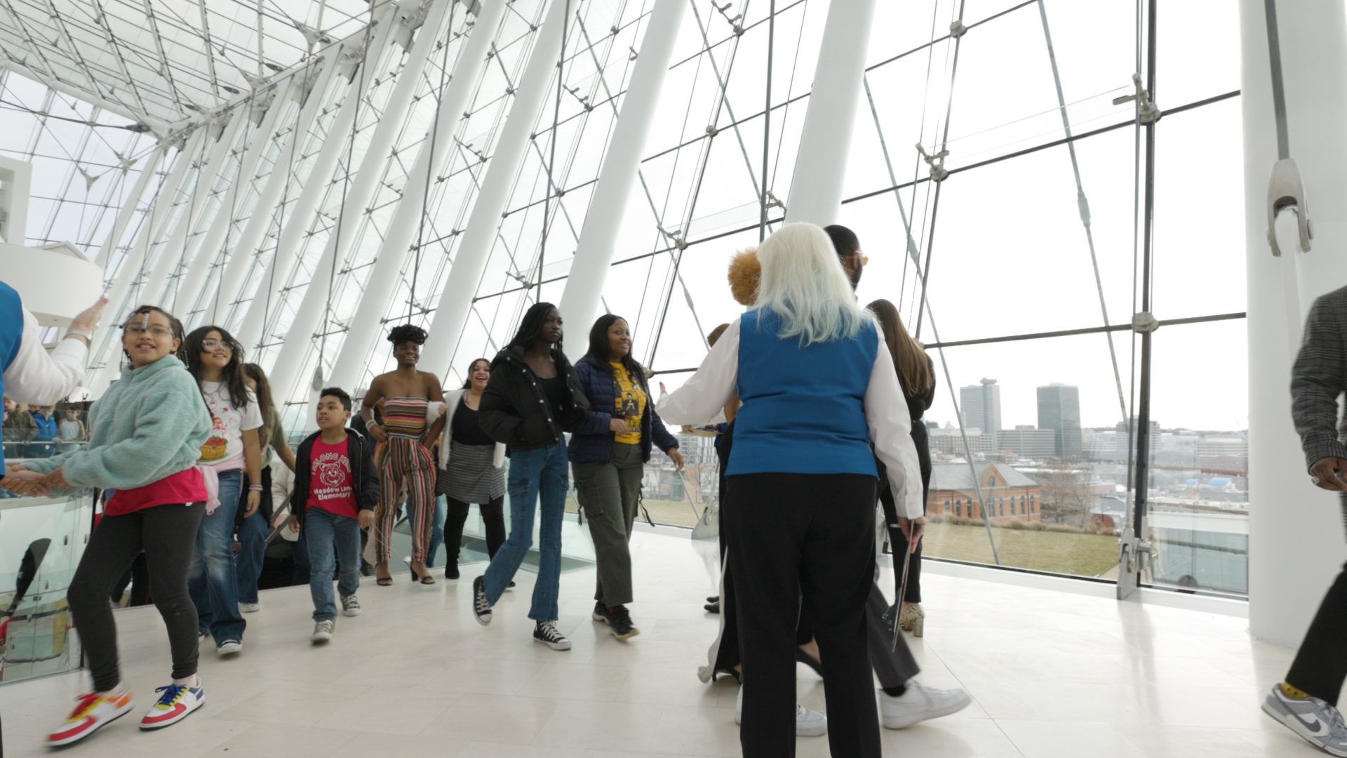 A group of people walk and interact inside the large, modern glass-walled Kansas City Performing Arts building, with a view of the cityscape in the background.
