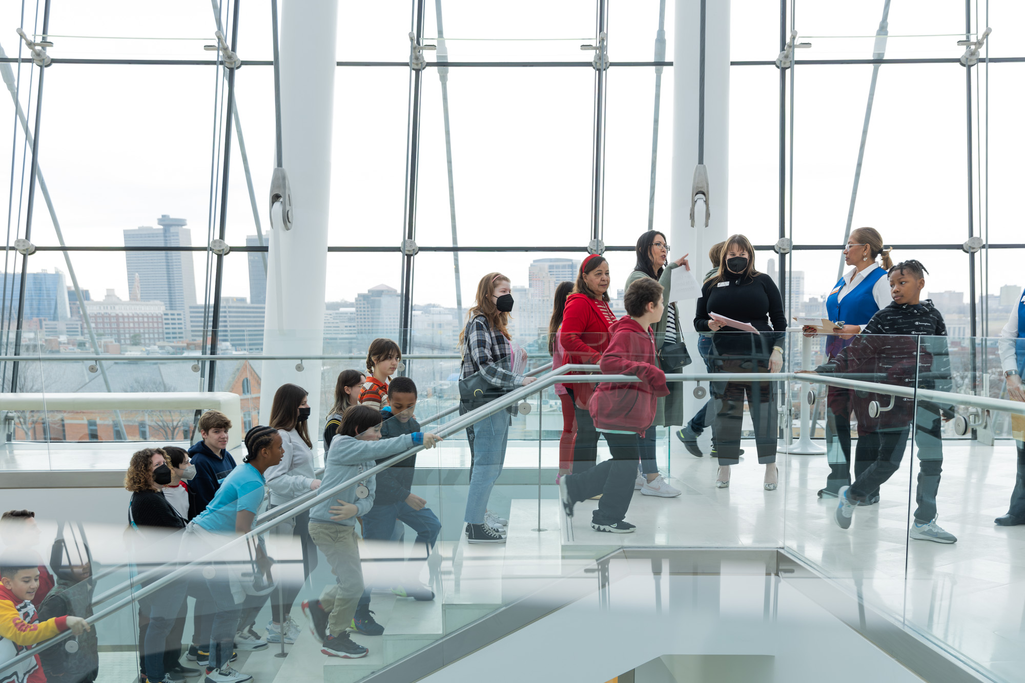A group of people, including children and adults, stands and walks on a modern glass staircase inside a building with large windows overlooking a cityscape.
