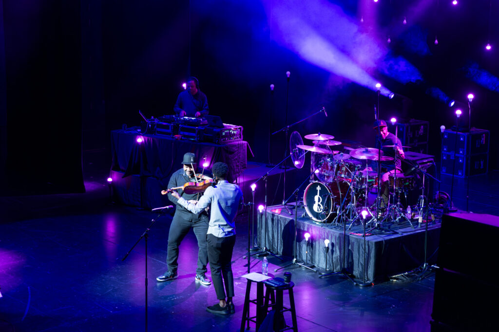 Musicians perform on stage with vibrant lighting: a violinist and a vocalist in the foreground, a drummer on a drum set to the right, and a DJ at a turntable in the background. This scene could be straight out of A Guide to the Kauffman Center.