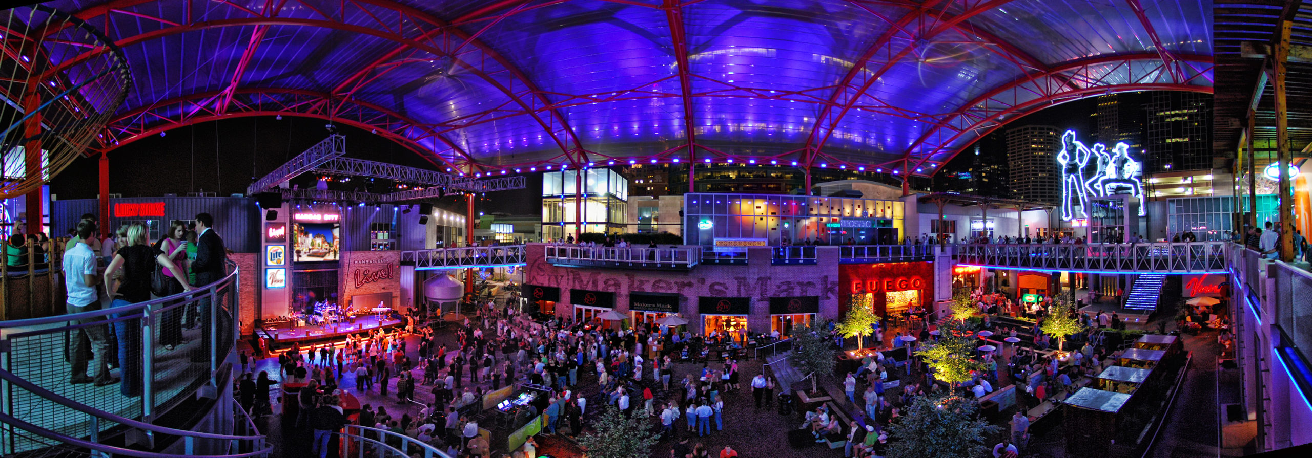 A vibrant indoor venue with a high roof is illuminated by blue and purple lighting, featuring a stage, large crowd, balcony seating, and restaurant spaces observed during a nighttime event.