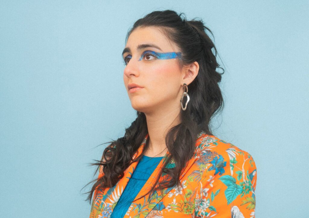 A person with long dark hair, wearing colorful makeup and a vibrant, patterned jacket, poses against a blue background.