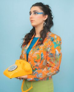 A woman with colorful eyeshadow holds a yellow rotary phone. She wears a vibrant orange jacket with a parrot print and stands against a light blue background.