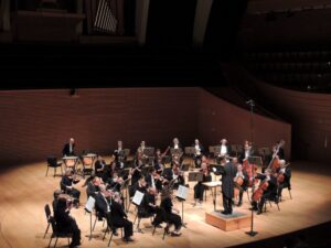 An orchestra performs on a concert hall stage with a conductor standing on a podium, surrounded by musicians playing various instruments.