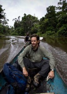 A man sits in a wooden canoe moving through a river in a jungle area. Another person is visible steering the canoe. The man has a backpack and is dressed in outdoor gear. Dense trees surround the river.