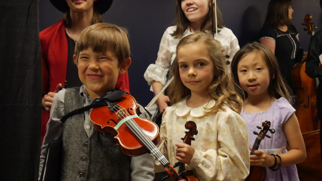 Young children holding violins, standing in a group, with some dressed in formal wear and others in casual clothing.