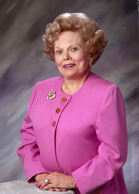 An older woman with curly hair stands facing the camera wearing a pink blazer and brooches. She is smiling and has her hands resting on a marble surface.