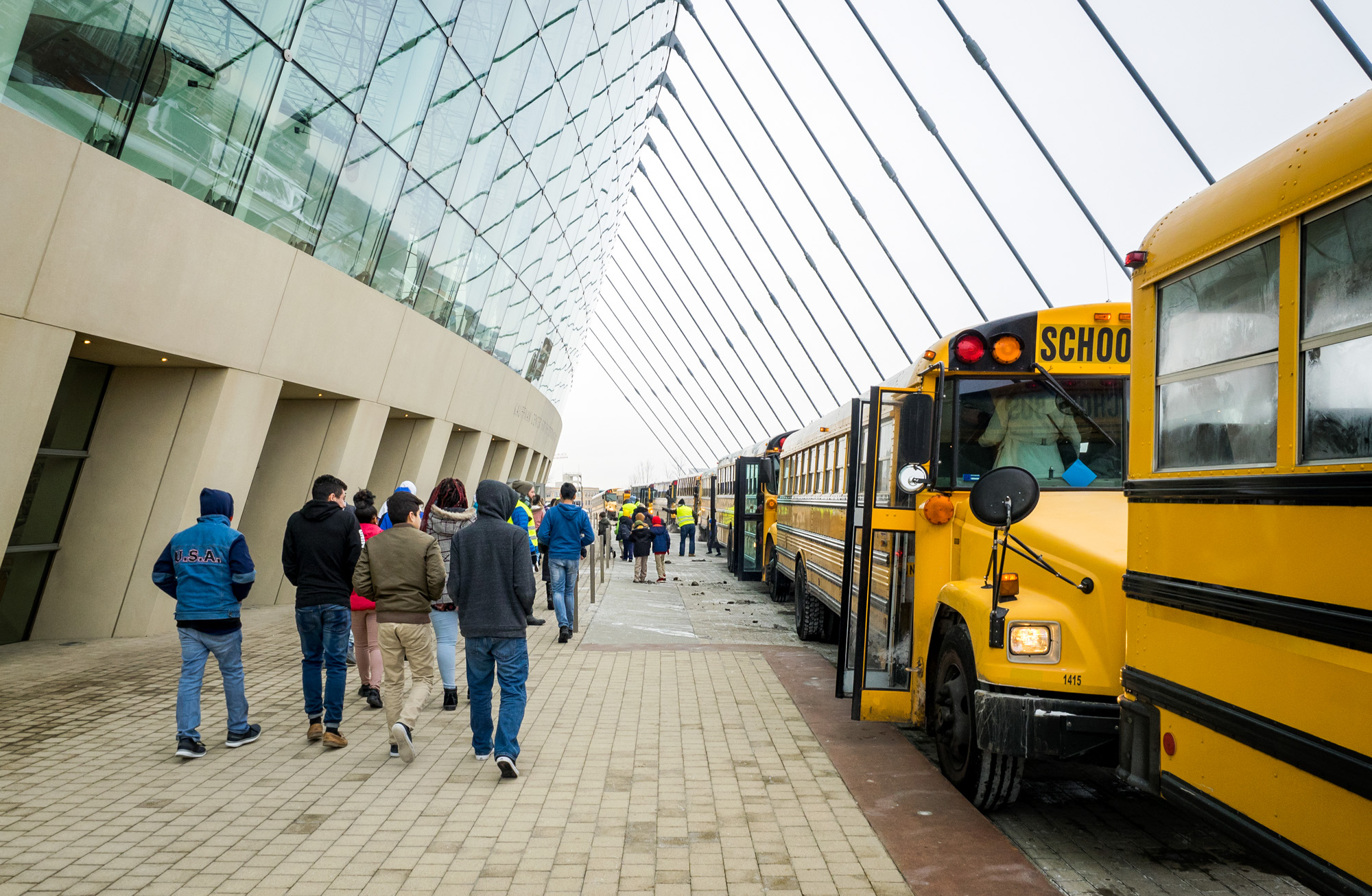 People walking on a paved path alongside parked school buses near the modern glass and concrete Kansas City Performing Arts building.