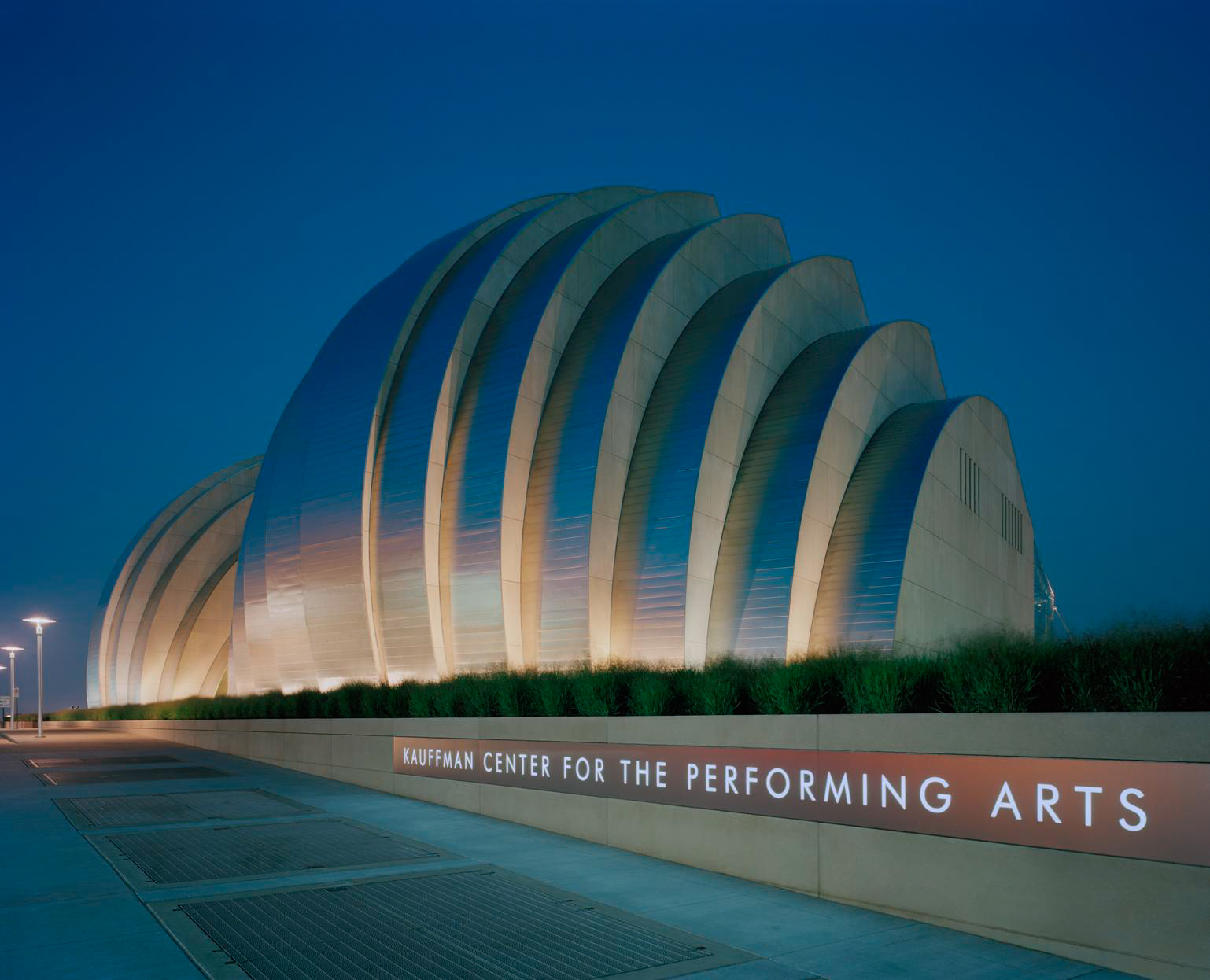 The image shows the Kauffman Center for the Performing Arts at night, featuring its distinctive curved architectural design, illuminated with soft lighting. This venue not only hosts world-class performances but also offers volunteer opportunities in Kansas City.