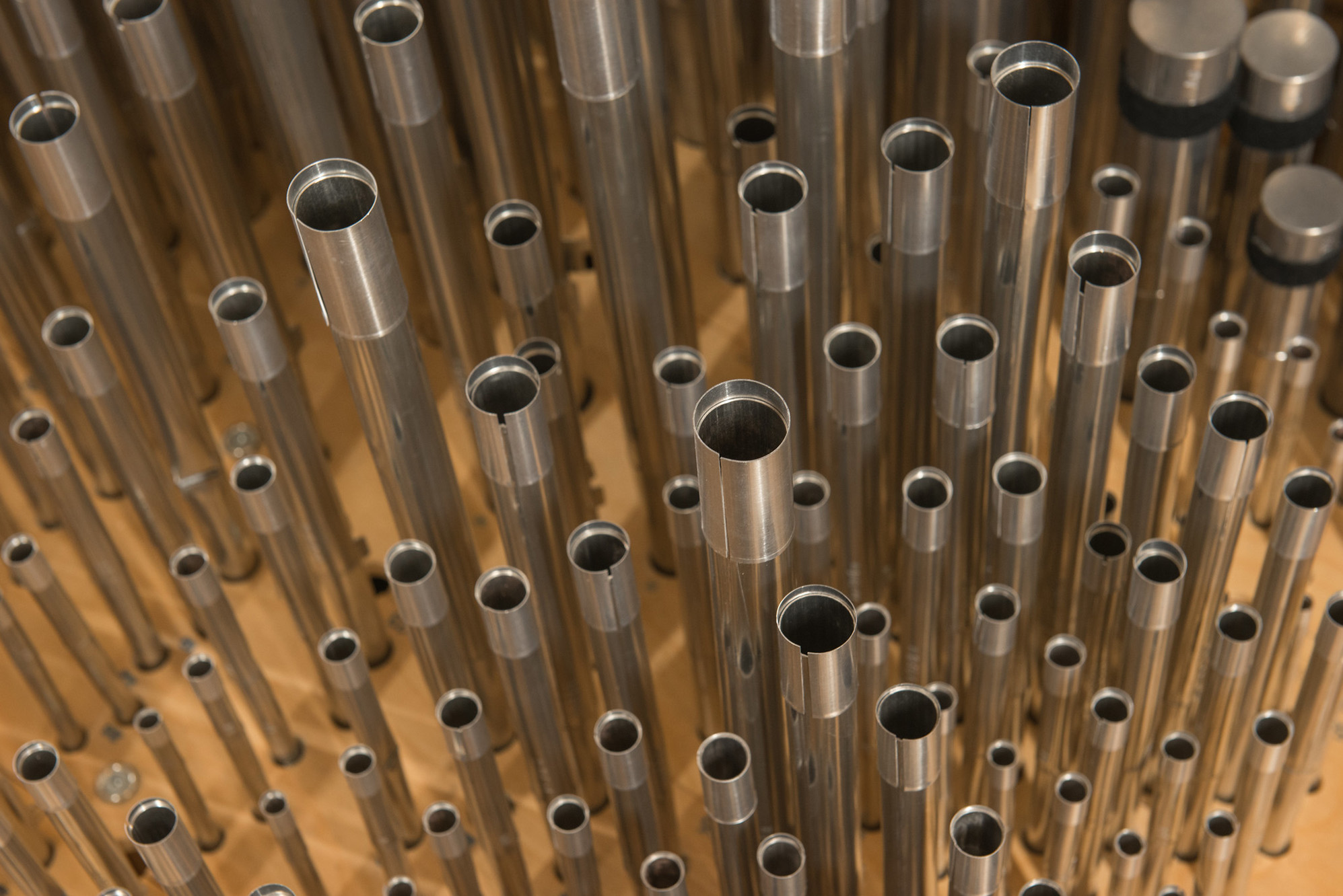 A close-up view showing an array of shiny, metal organ pipes of varying lengths and diameters, tightly clustered together and extending vertically in different directions. The background shows a wooden surface, adding a warm contrast to the metallic appearance of the Julia Irene Dennie-Kauffman Casavant Organ.