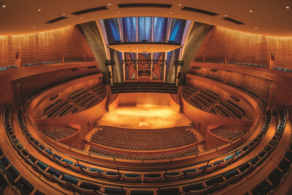 A wide-angle view of an empty, modern concert hall with multiple levels of seating, wooden interiors, and a well-lit stage.
