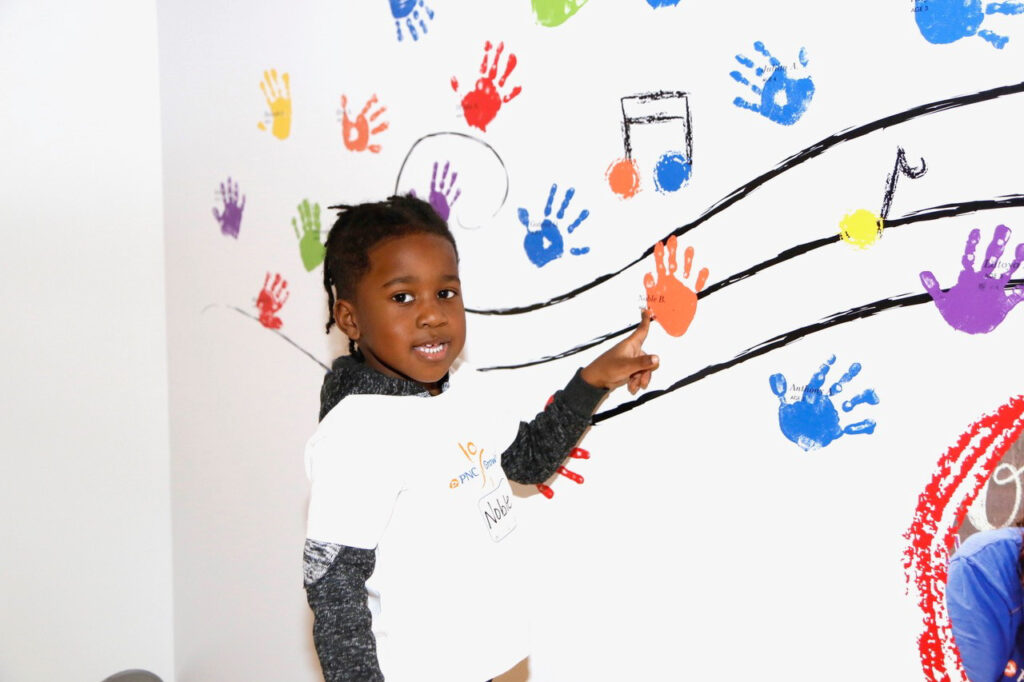 A child points at colorful handprints and musical notes painted on a white wall.