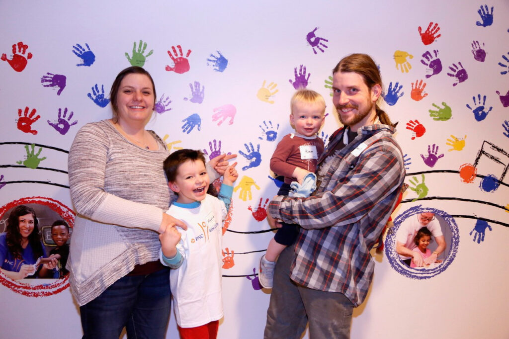 A smiling family of four stands in front of a colorful handprint mural. The parents hold their two young children, with the older child in front and younger child being held by the father.