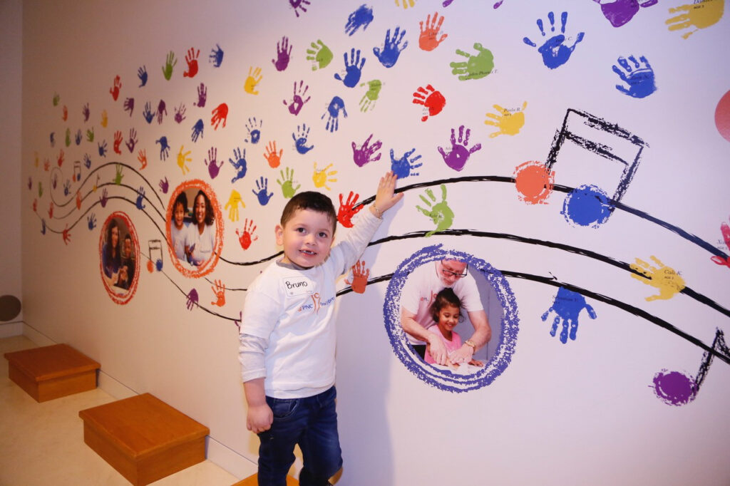 A child stands in front of a colorful wall with handprints and musical notes, touching the artwork. Brightly colored handprints and photos of children decorate the wall.