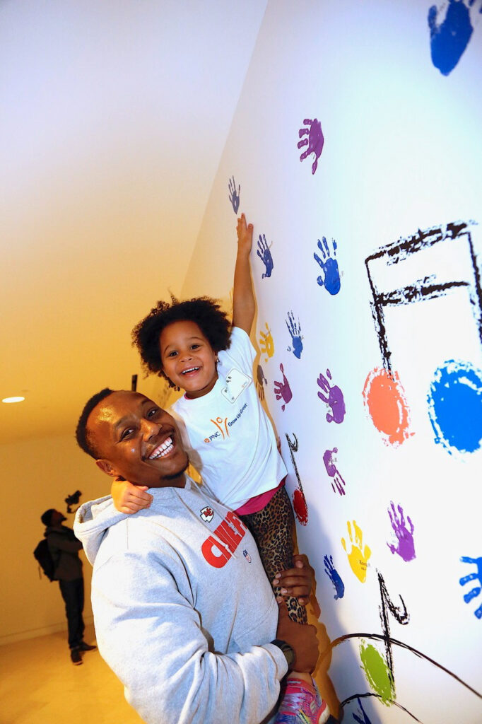 A smiling man lifts a child who is reaching to touch colorful handprints on a white wall.