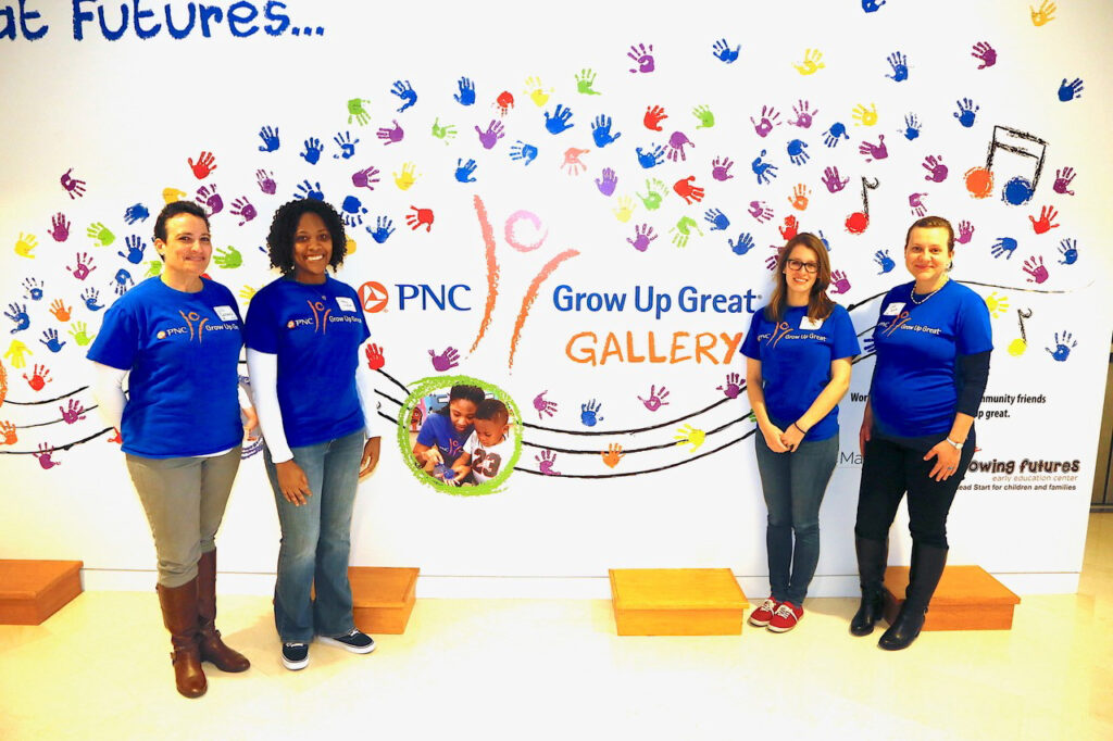 Four women wearing blue shirts stand in front of a colorful wall with handprints and the PNC Grow Up Great Gallery logo.