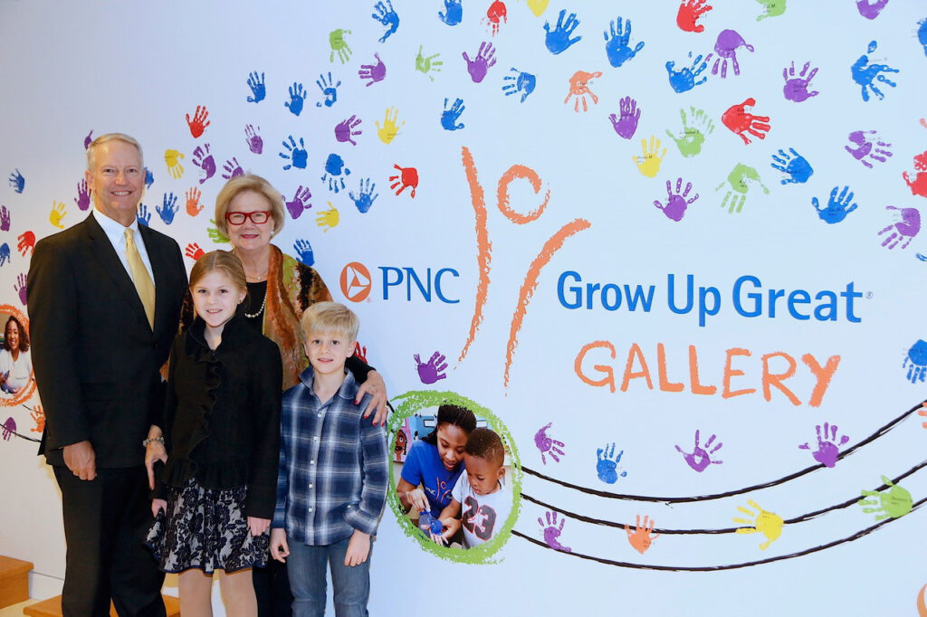 A man, a woman, and two children stand in front of the PNC Grow Up Great Gallery wall, which is decorated with colorful handprints.