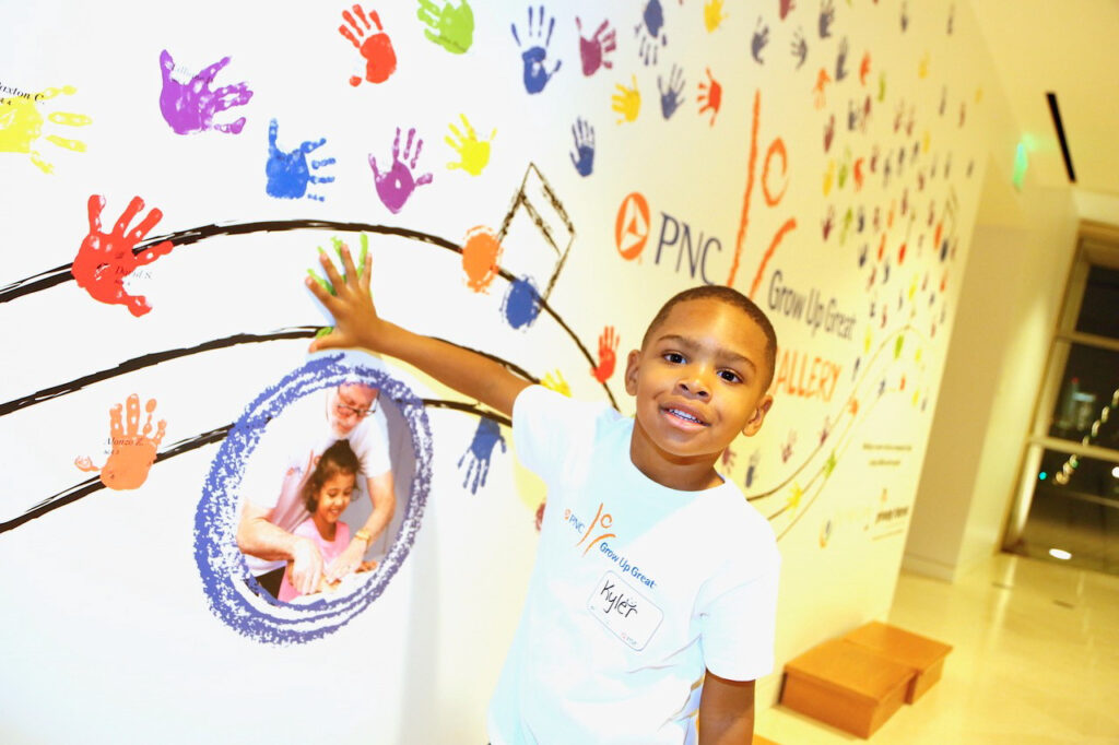 A child pressing a painted handprint on a mural adorned with colorful handprints. The wall, beautifully branded as the PNC Grow Up Great Gallery, features a circular photo of two people. The child wears a white shirt.