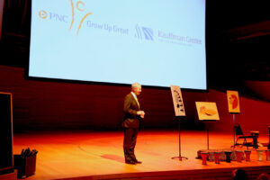 A person in a suit stands on a stage next to three easels holding art pieces. Behind them, a screen displays logos for PNC Grow Up Great and Kauffman Center for the Performing Arts.