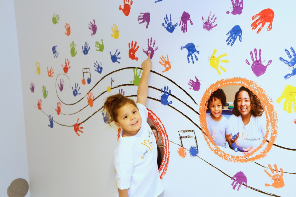 Child points to a wall with multicolored handprints and musical notes. Two circular images featuring people are incorporated into the mural.