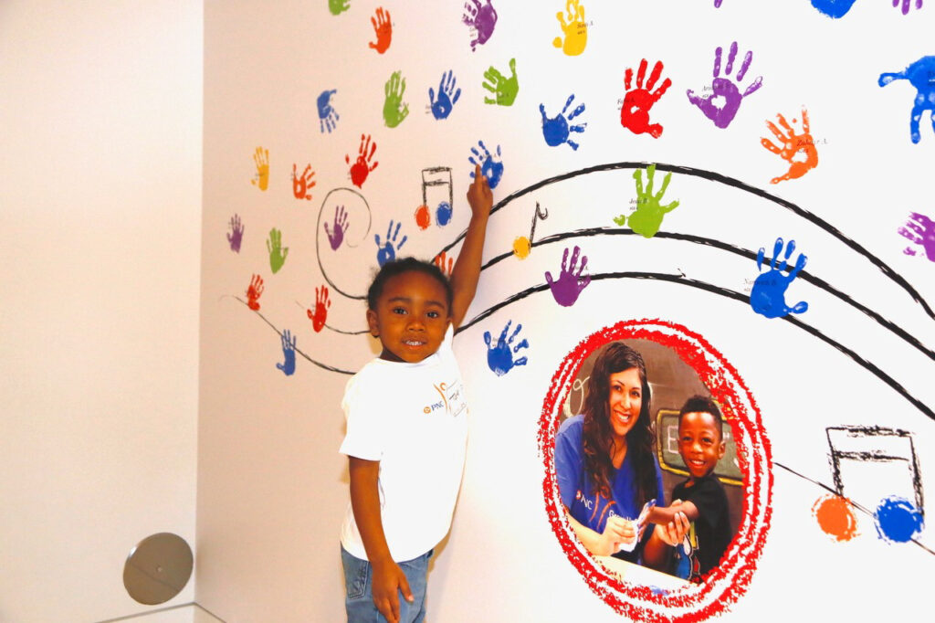 Child pointing to colorful handprints on a wall mural with a photo of a smiling adult and child inside a red circle. Musical notes are part of the mural design.