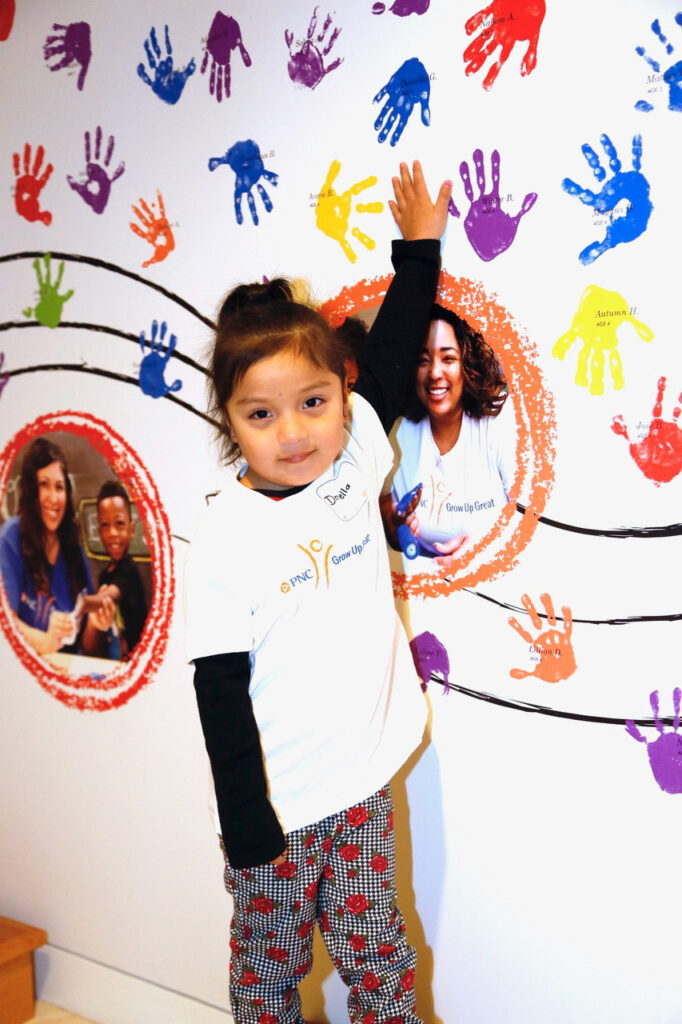 A child wearing a white shirt stands in front of a wall decorated with colorful handprints and circular cutouts of people, reaching up to touch one of the handprints.