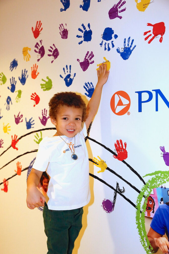 A smiling child stands in front of a wall covered in colorful handprints, reaching up to add a yellow handprint. He is wearing a white shirt with a design on it.