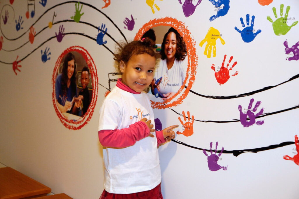 A young girl in a white shirt points at colorful handprints on a wall mural. The mural also features circular photos of other people and more handprints in various colors.