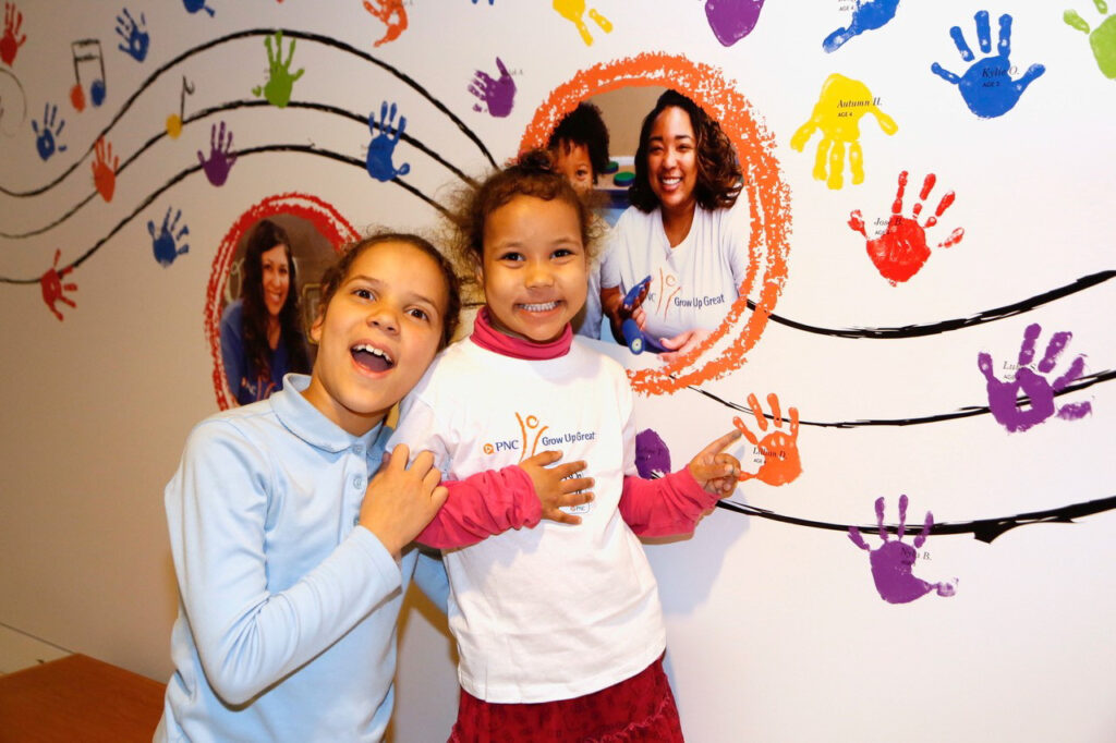 Two children stand smiling in front of a wall decorated with colorful handprints and circular photos of people. One child is wearing a white PNC t-shirt and the other is in a light blue sweater.