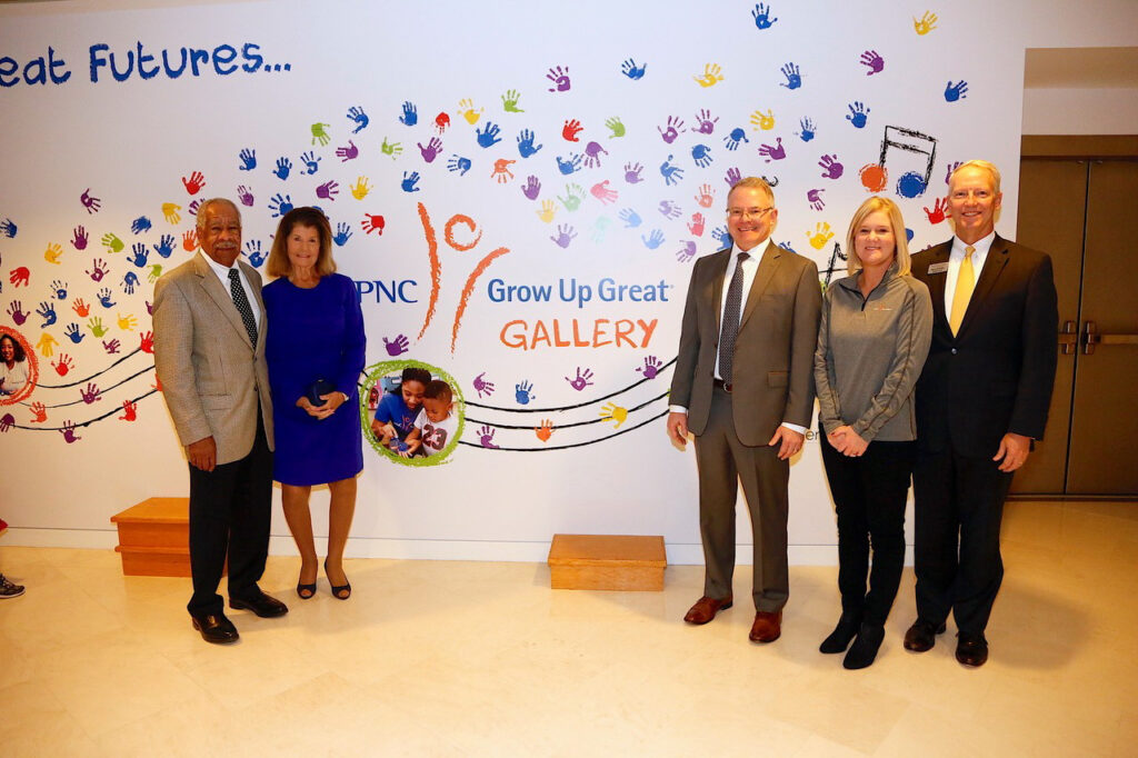 Five people stand in front of a wall with colorful handprints and the text "PNC Grow Up Great Gallery.