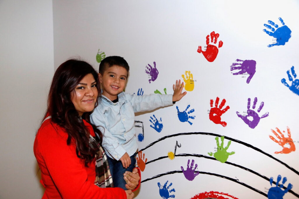 A woman in a red shirt holds a young boy who is touching colorful handprints on a white wall.