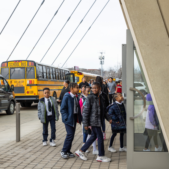 Children are entering a building from a patio area while two yellow school buses are parked in the background.