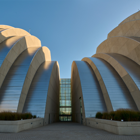 A modern architectural building featuring rounded, ribbed silver and beige structures with a glass entrance in the center, viewed from the ground level against a clear blue sky.