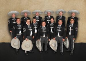 A mariachi band poses for a group photo, all dressed in traditional black charro suits with silver embroidery, wide-brimmed sombreros in hand. There are twelve members standing against a plain backdrop.