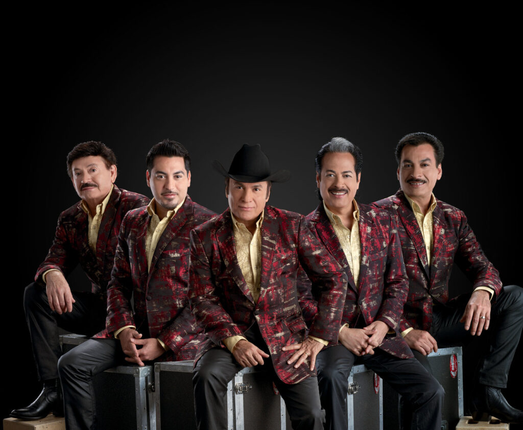 Five men are seated and wearing matching red and gold jackets, with one man in the center also wearing a black cowboy hat. They are posing against a black background.
