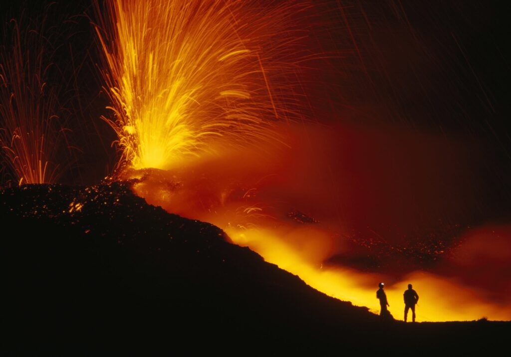 Two people stand observing a dramatic volcanic eruption at night, with bright orange lava and sparks contrasting against the dark sky.