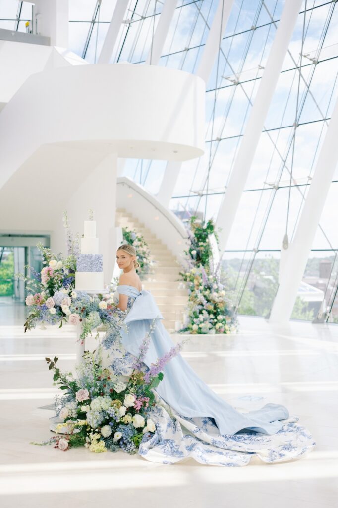 A woman in a light blue, flowing dress stands by a multi-tiered white cake adorned with flowers in a modern, glass-walled building with a grand staircase and floral decorations.