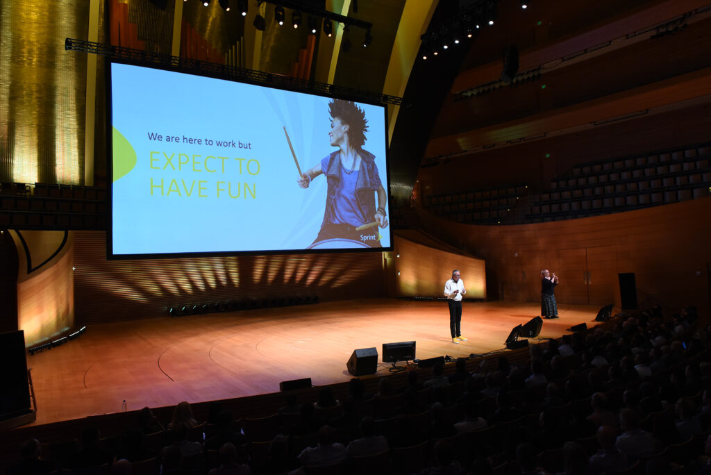 A speaker stands on a stage with a large screen behind them displaying the text: "We are here to work but EXPECT TO HAVE FUN." An audience is seated in the spacious Kauffman Center auditorium, available for rent.