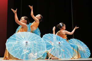 Three performers in traditional attire hold painted blue umbrellas, kneeling with raised arms during a dance on stage.