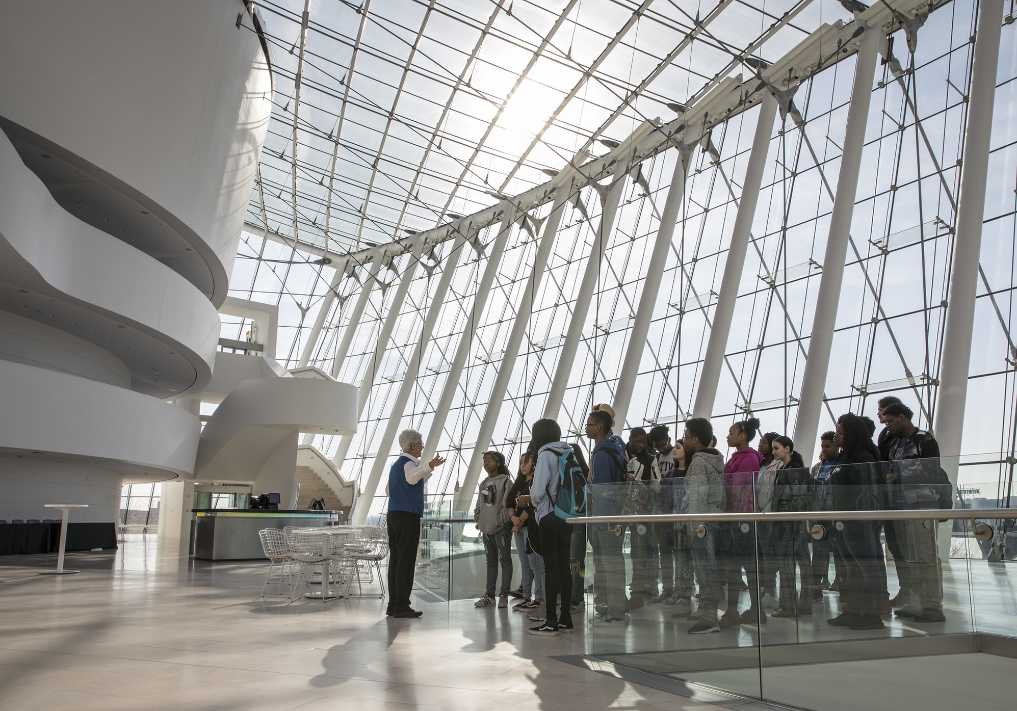 A group of people stands attentively while an older person gestures and speaks to them in a spacious, modern building with large, sunlit glass windows and a curved architectural design. The interior features sleek white surfaces and a few tables and chairs, perfect for educational Field Trips in Kansas City.