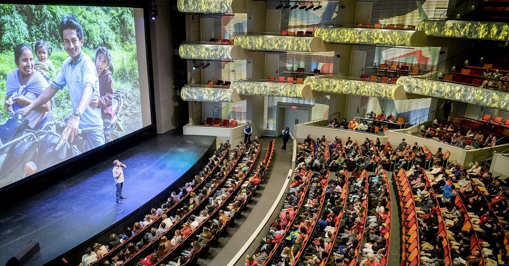 A large proscenium theater filled with audience members watching a presentation with a speaker and large screen featuring photography.
