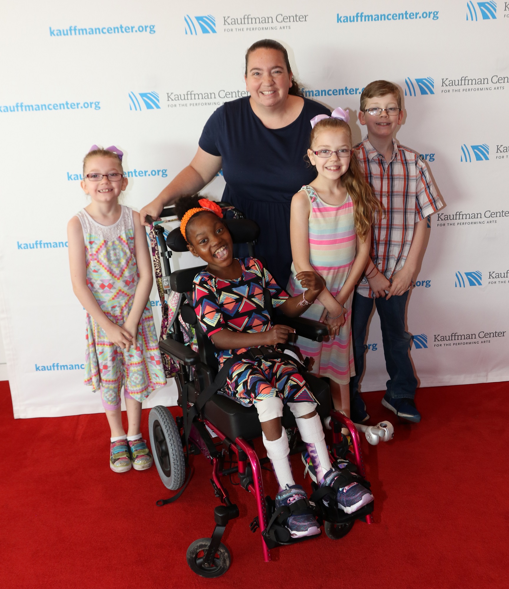 A group of five people, including a standing woman, three standing children, and one child using a wheelchair, pose for a photo against a white backdrop with the blue Kauffman Center logo repeated.