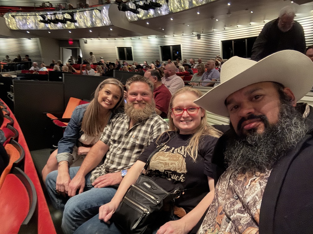 In the foreground, four seated patrons pose for a selfie inside of Muriel Kauffman Theatre. The photographer wears a cowboy hat and camouflage shirt. In the background, dozens of other patrons take their seats in the red and orange theater seats under the silver balustrades. 