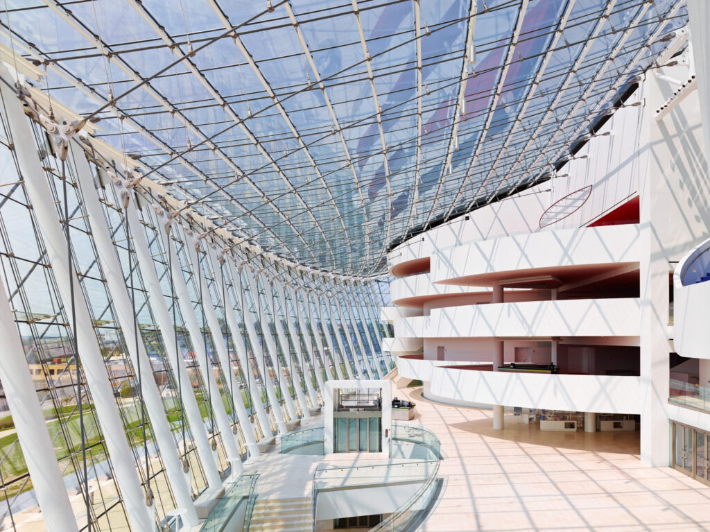 Modern indoor space featuring a large glass ceiling and walls, white structural elements, and multiple balconies. The expansive design allows for abundant natural light.