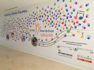 A colorful wall mural with handprints, photos, musical notes, and logos of PNC, St. Mark Center, Metro Center, Growing Futures, and El Centro, promoting a program for children's growth and education.