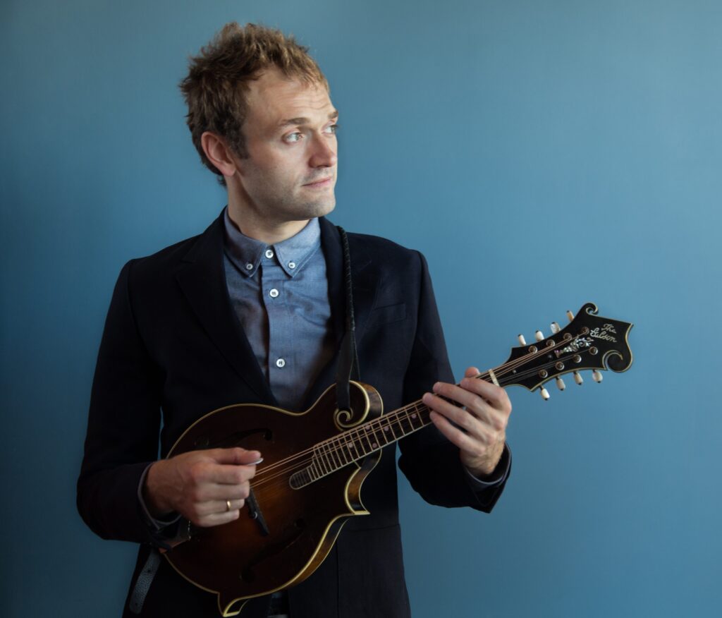 A person in a suit stands against a blue background, holding and playing a mandolin.