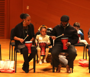 A seated family of three plays hand drums with mallets. The child in the center is smiling between the adults on either side in a warmly lit room.