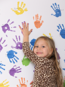 A young girl in a leopard-print top places her hand on a wall filled with colorful handprints.